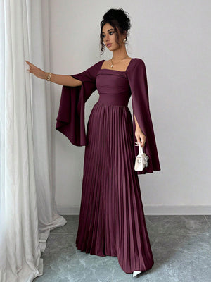 A woman models a floor-length plum purple pleated gown with a square neckline, a fitted bodice, and dramatic draped cape sleeves. She is holding a small white handbag and posing in front of a white curtain and gray wall.