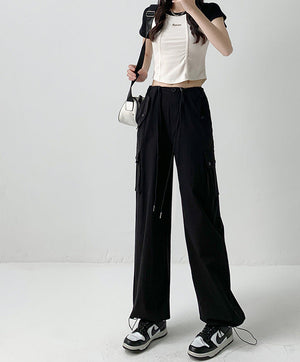A model wearing black wide-leg cargo pants with a drawstring waist, paired with a white ribbed crop top and black-and-white sneakers, standing against a light gray studio background. Chinese text overlays are present on the right side.