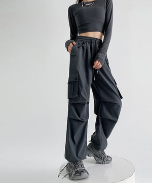 A model wearing dark gray wide-leg cargo pants with a drawstring waist, paired with a matching long-sleeve crop top and gray sneakers, standing on a white circular platform against a light gray background. Chinese text overlays are present on the right side.