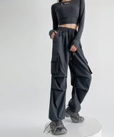 A model wearing dark gray wide-leg cargo pants with a drawstring waist, paired with a matching long-sleeve crop top and gray sneakers, standing on a white circular platform against a light gray background. Chinese text overlays are present on the right side.
