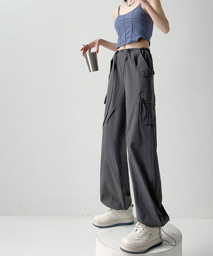 A model wearing dark gray wide-leg cargo pants with a drawstring waist, styled with a blue ribbed crop top and white sneakers, standing on a white pedestal against a light gray studio background. Chinese text overlays are present on the right side.